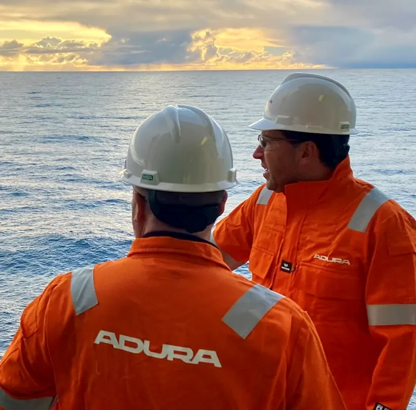 Two workers in orange safety uniforms and white hard hats stand on an offshore installation, looking out over the ocean
