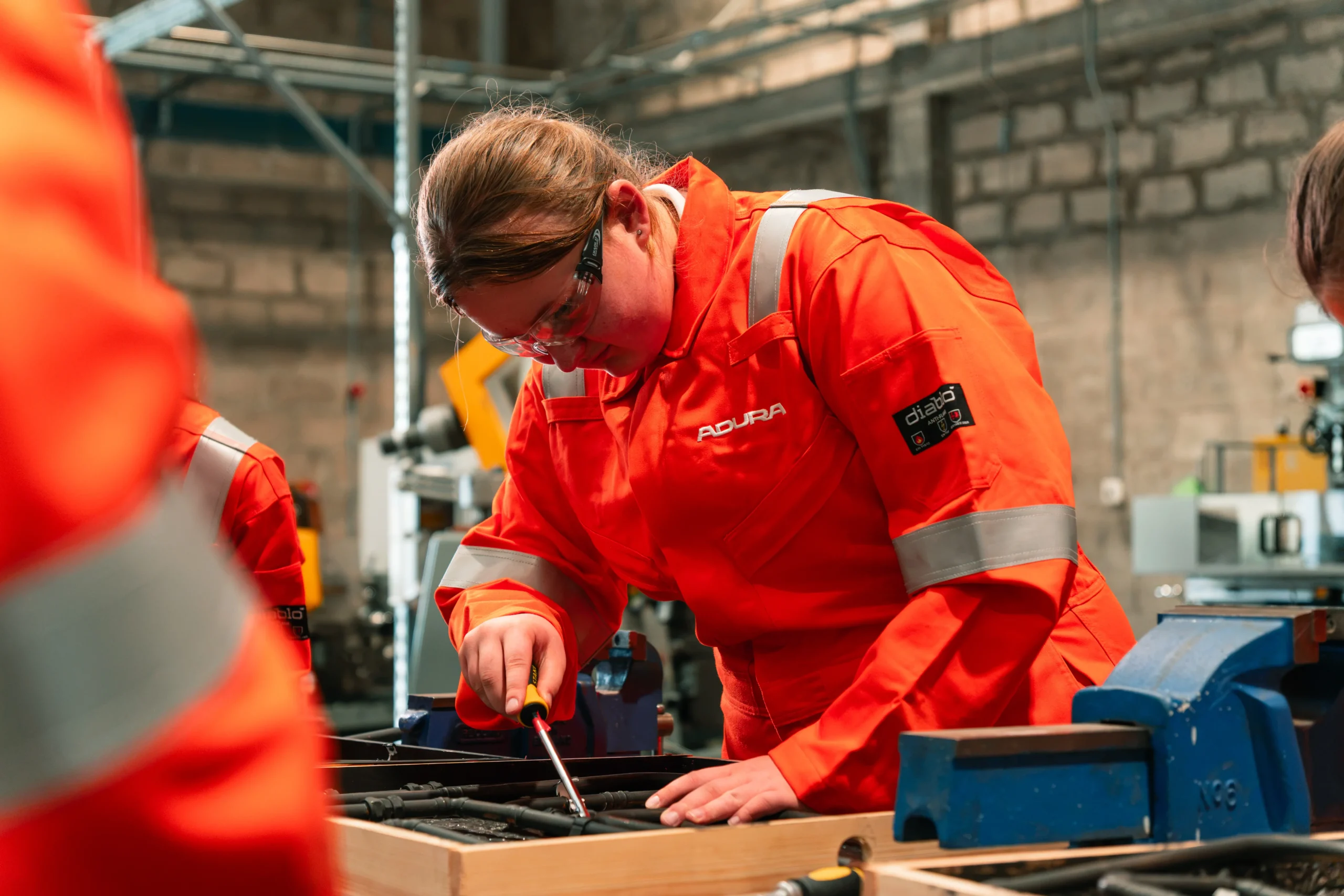 A Girls in Energy student in orange Adura coveralls and safety glasses using a screwdriver on equipment at a workshop bench
