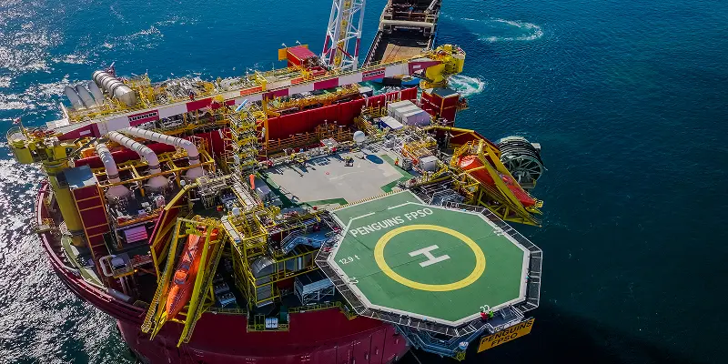 Aerial view of an offshore oil platform, featuring a green helipad labeled "Penguins FPSO." Surrounded by blue ocean, the structure displays complex machinery and pipes
